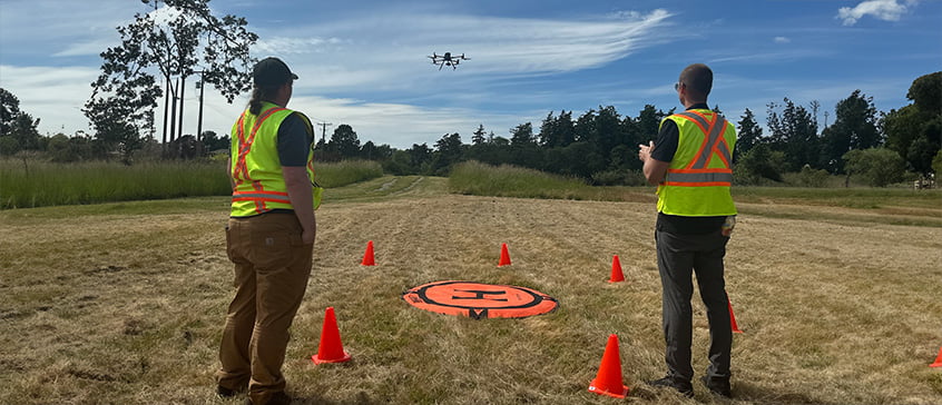 Two people in high-visibility vests operate a drone in an open grassy field. The drone is flying above, and an orange landing pad with a black "H" is on the ground, surrounded by four orange traffic cones. Trees and a partly cloudy blue sky are in the background.