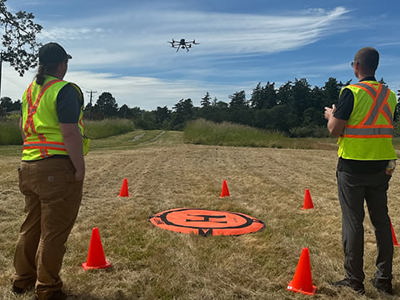 Two people in high-visibility vests operate a drone in an open grassy field. The drone is flying above, and an orange landing pad with a black "H" is on the ground, surrounded by four orange traffic cones. Trees and a partly cloudy blue sky are in the background.