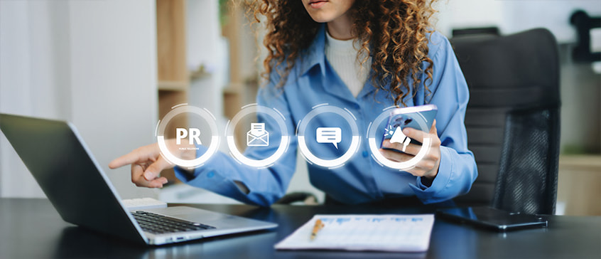 A woman with curly hair wearing a blue shirt sits at a modern office desk working on a laptop, with documents nearby. Four translucent circular icons float above the workspace displaying 