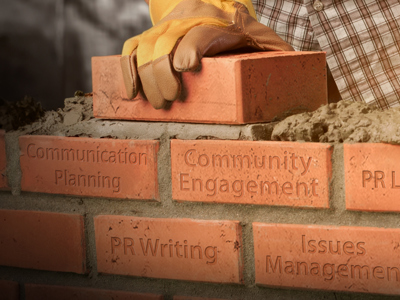 A close-up of a brick wall being built, with a gloved hand placing a brick and a trowel adding mortar. Each brick is etched with a public relations concept: Communication Planning, Community Engagement, PR Leadership, PR Writing, Issues Management, and Media Relations. This image visually represents building foundational skills for a successful PR career.