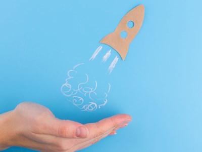 A hand beneath a sketched rocket ship taking off against a light blue background.