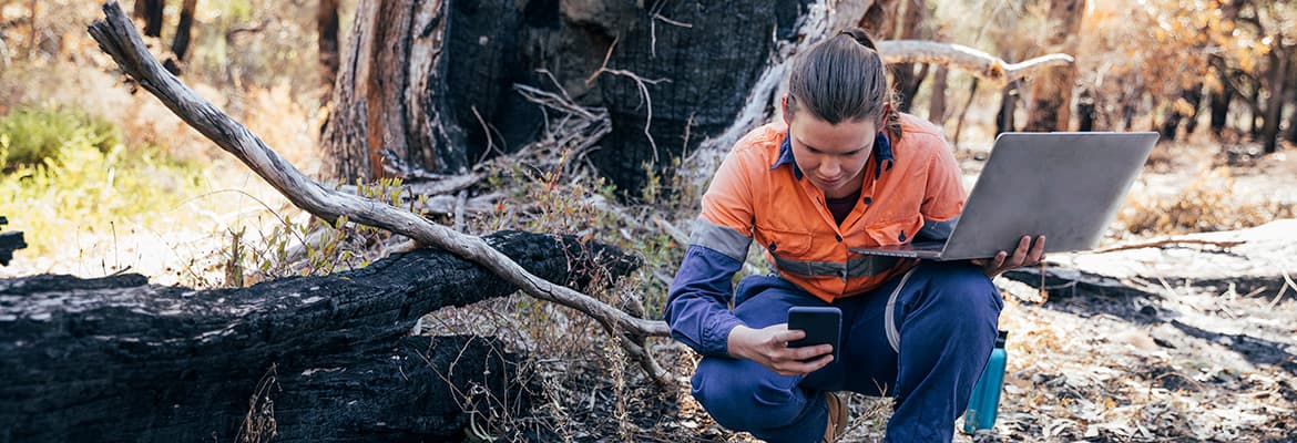 Student crouching down in a dry forest area taking samples and working on a laptop.