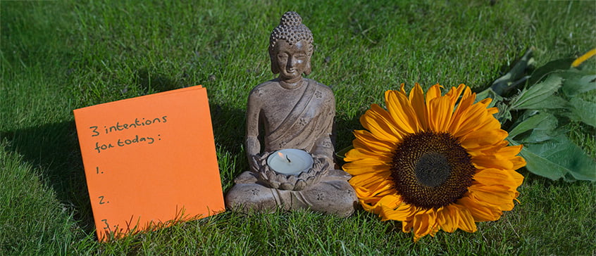 Buddha statue on the grass with burning candle, a sunflower and the awareness of 3 intentions for today.
