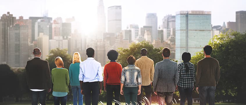 A group of people standing in a line with their backs towards the camera as they look over a city skyline in the sunshine. 