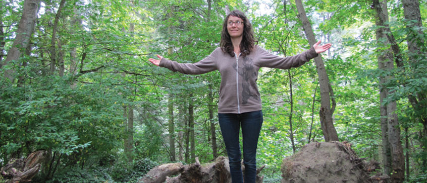 Amanda Evens wearing a grey hoodie with a grizzly bear graphic stands, arms wide in a lush green forest.