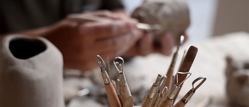 Hands shaping a clay sculpture with pottery tools in the foreground.