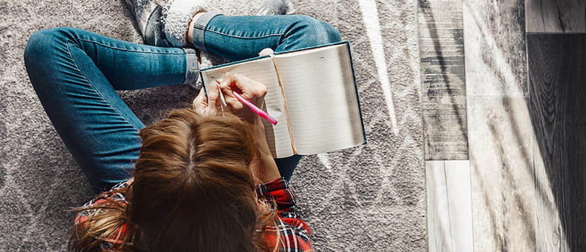 Overhead view of a person with brown hair lying on a textured rug, wearing jeans and a patterned top, writing in an open notebook with a pink pen, illustrating the personal and intimate nature of narrative writing and storytelling.