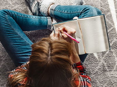 Overhead view of a person with brown hair lying on a textured rug, wearing jeans and a patterned top, writing in an open notebook with a pink pen, illustrating the personal and intimate nature of narrative writing and storytelling.