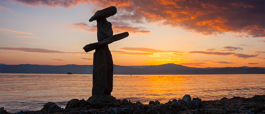 A peaceful sunset over calm water with mountains in the background. In the foreground, balanced stones are stacked in an Inukshuk formation on a rocky shoreline, with the sky glowing in shades of orange, pink, and purple.