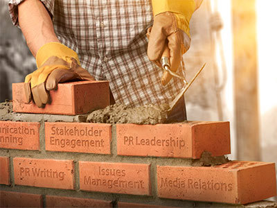 Construction worker layering bricks which have the following phrases engraved on them: stakeholder engagement, PR leadership, PR writing, issues management and media relations.