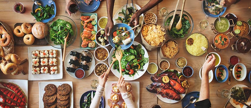 Overhead view of a dining table laden with a variety of dishes. Hands serving portions