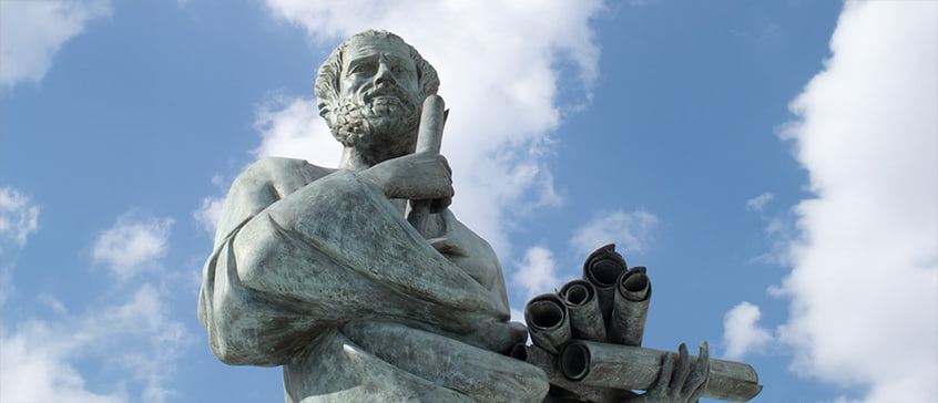 Statue of Aristotle holding scrolls against a blue sky, symbolizing classical philosophy and the study of virtue ethics in ancient Greek thought.