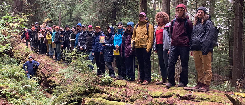 Large group of people wearing colourful rain jackets and outdoor gear standing together large fallen fir tree surrounded by moss-covered ground, ferns, and tall coniferous trees.