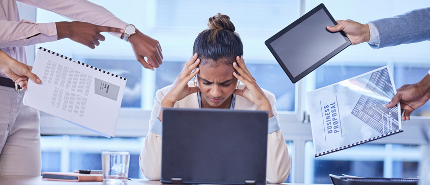 Woman at laptop with hands on forehead while four sets of hands are demanding reports to be done