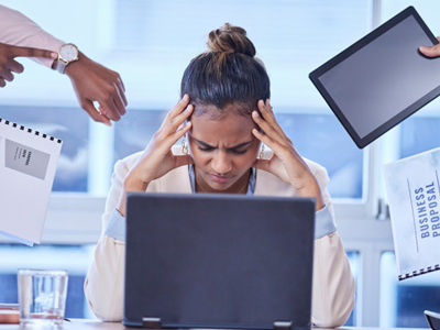 Woman at laptop with hands on forehead while four sets of hands are demanding reports to be done