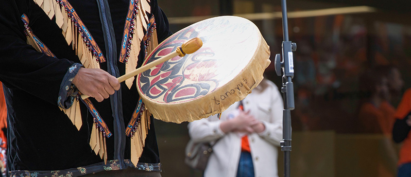 Close up shot of a person in a decorated black-coloured Indigenous jacket with beadwork and cedar fringes falling diagonally inward from the shoulders down. They are beating a hand drum with Coast Salish or Haida artwork on the skin. An Indigenous ceremony.  