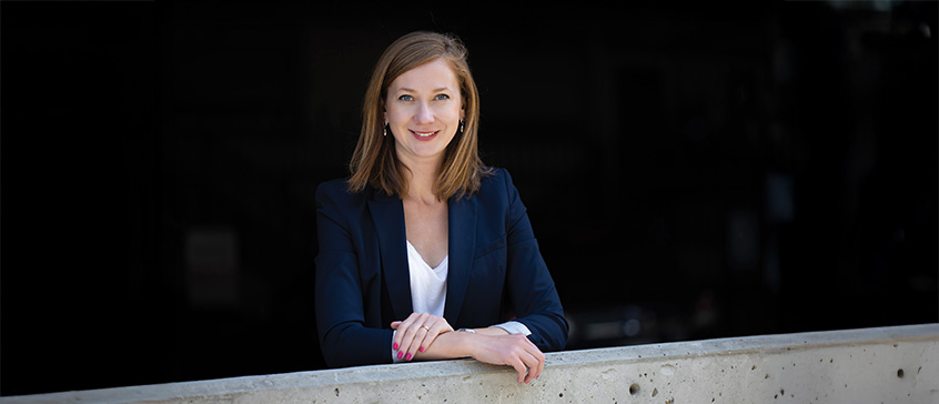 Galina Sanaeva, a professional educator, stands outdoors against a dark background, leaning on a concrete ledge. She is dressed in a navy blue blazer over a white top, with shoulder-length light brown hair and a clear, confident expression.
