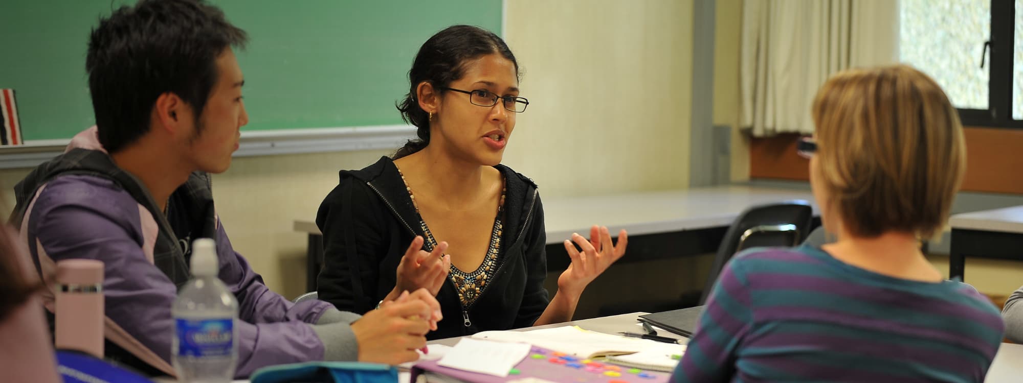 Three people engaged in discussion around a classroom table, with papers and notebooks in front of them.
