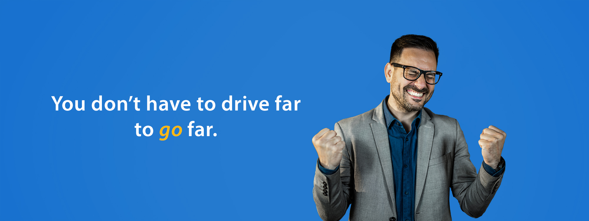 A man in a blazer and glasses smiles and pumps his fists in excitement against a solid blue background. Beside him, white and yellow text reads: “You don’t have to drive far to go far.”