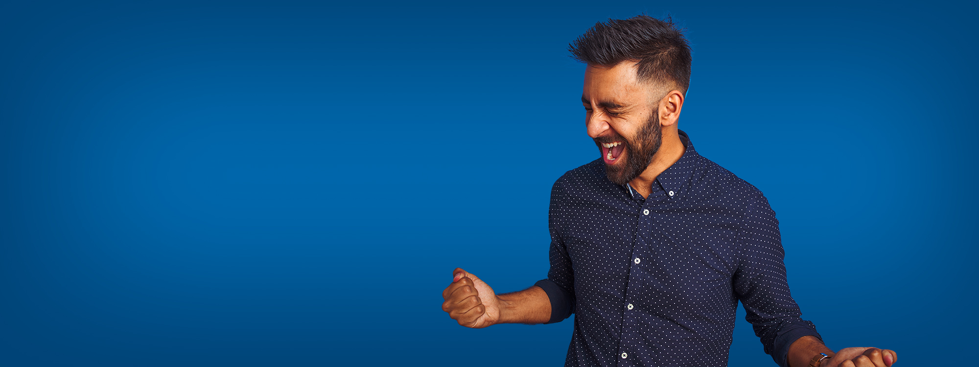 A smiling man with a beard wearing a navy patterned button-up shirt, celebrating with a clenched fist against a blue background.