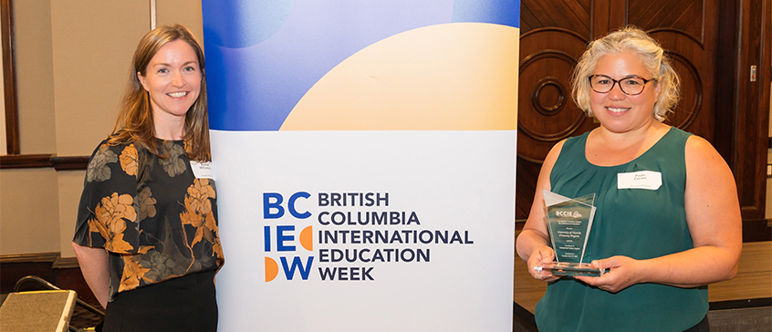 Two women standing beside a banner that reads “British Columbia International Education Week.” One woman is smiling at the camera, and the other is holding an award.