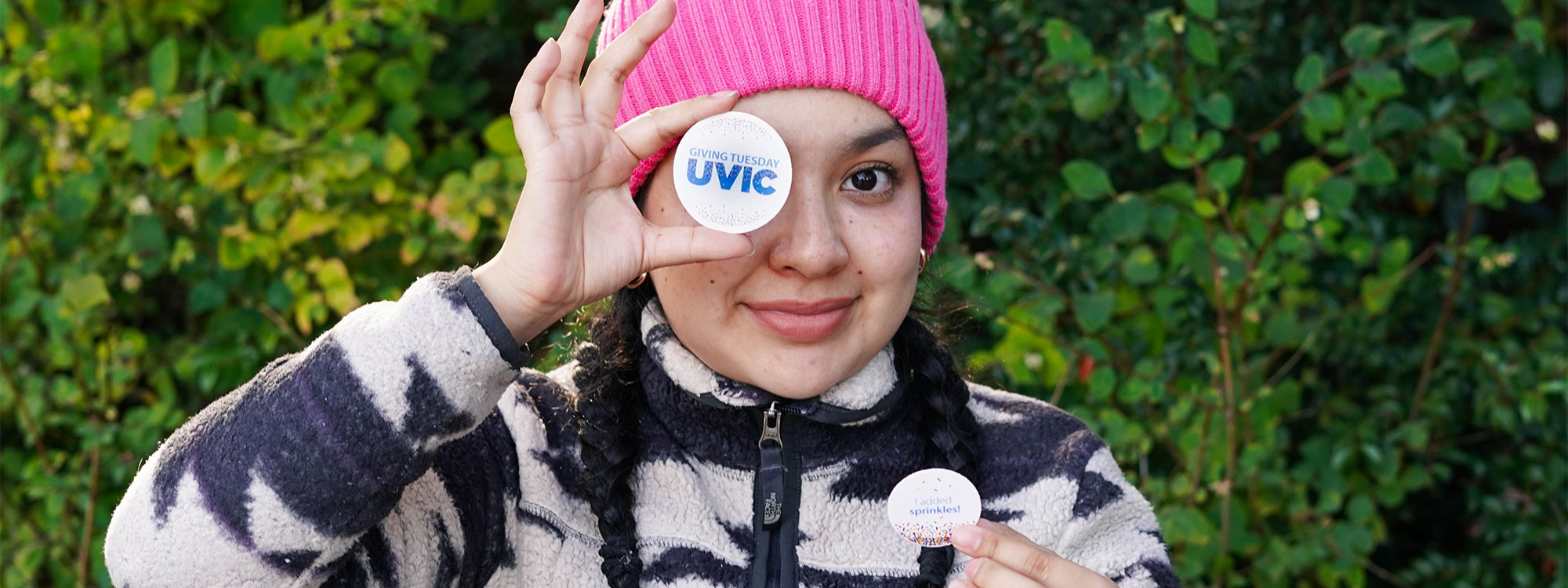 A young woman wearing a pink beanie holding up a UVic sticker.