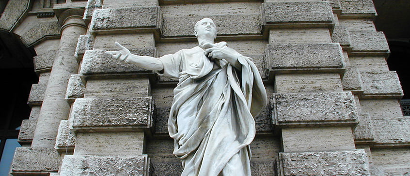 A full-length white marble statue of Roman orator, Cicero stands before a textured stone wall. The figure is draped in flowing robes, with one arm outstretched in a gesturing pose and the other holding scrolls or papers against its chest. The statue