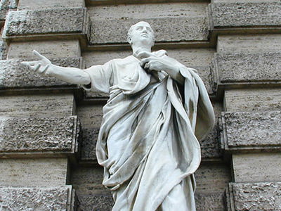  A full-length white marble statue of Roman orator, Cicero stands before a textured stone wall. The figure is draped in flowing robes, with one arm outstretched in a gesturing pose and the other holding scrolls or papers against its chest. The statue