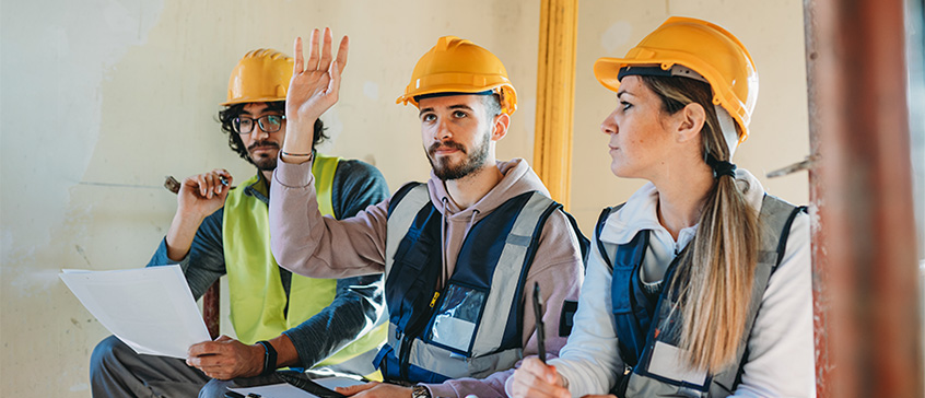 Construction worker raising his hand with a question while sitting beside two other construction workers. 