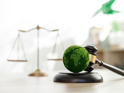 A gavel rests beside a green globe covered in grass, with a justice scale in the background.