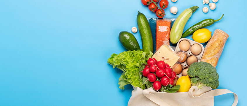 A cloth grocery bag lying on a blue background with produce and other food spilling out.