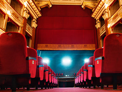 Interior view of an ornate theatre with rows of red velvet seats, illuminated stage, and gold architectural details in the balconies