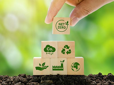 A hand stacks wooden blocks with eco-friendly icons on soil, highlighting net zero emissions against a green background.