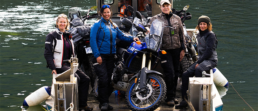 Suzanne and 3 other riders, smiling and standing on a small boat with their motorcycles, green water surrounds them. 