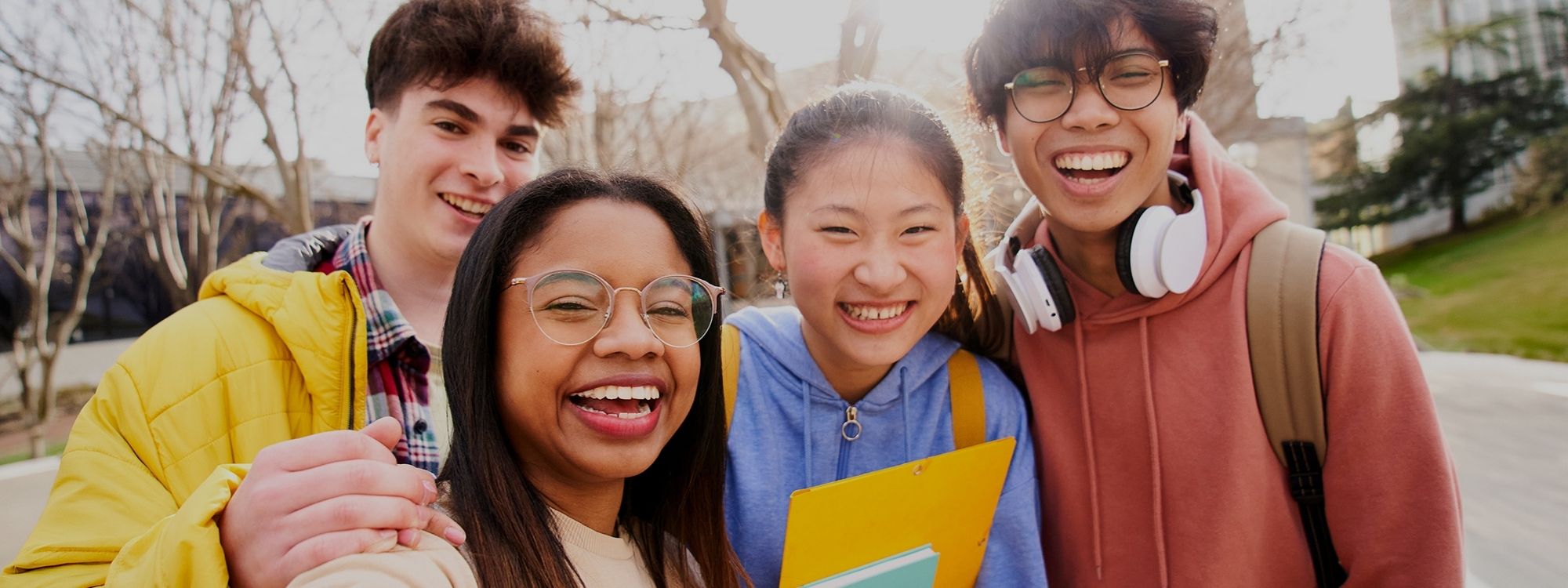 Four smiling teenagers take a selfie together outdoors on a sunny campus, one holding notebooks.