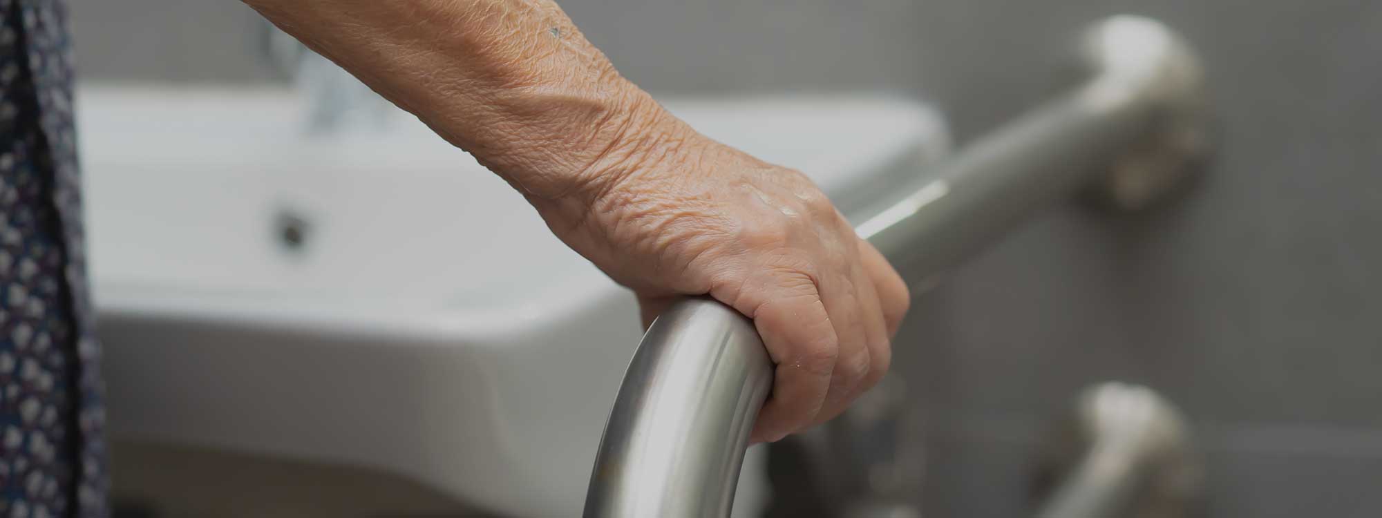 An older person's hand holds onto a wall-mounted grab bar in a bathroom, aiding balance and fall prevention.