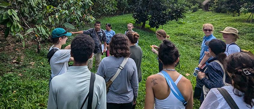 A diverse group of students and instructors from the UVic Agroecology Field School in Belize gathered in a circle on lush green grass, engaged in an outdoor learning discussion. Participants wear casual field clothing and backpacks, with dense tropical vegetation forming a natural classroom backdrop.