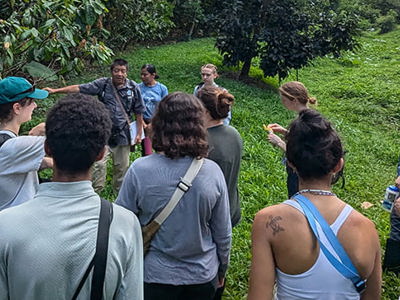 A diverse group of students and instructors from the UVic Agroecology Field School in Belize gathered in a circle on lush green grass, engaged in an outdoor learning discussion. Participants wear casual field clothing and backpacks, with dense tropical vegetation forming a natural classroom backdrop.