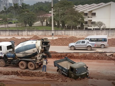 Construction site with concrete mixer trucks and dump trucks on dusty, unpaved ground. Workers visible near heavy machinery without clear protective equipment. Residential buildings nearby suggest potential community exposure to construction dust and noise pollution, impacting both worker respiratory health and air quality for local residents.