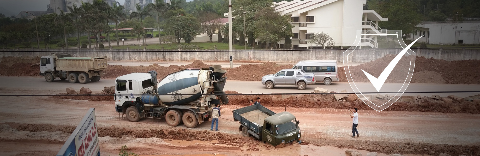 Construction site with concrete mixer trucks and dump trucks on dusty, unpaved ground. Workers visible near heavy machinery without clear protective equipment. Residential buildings nearby suggest potential community exposure to construction dust and noise pollution, impacting both worker respiratory health and air quality for local residents.