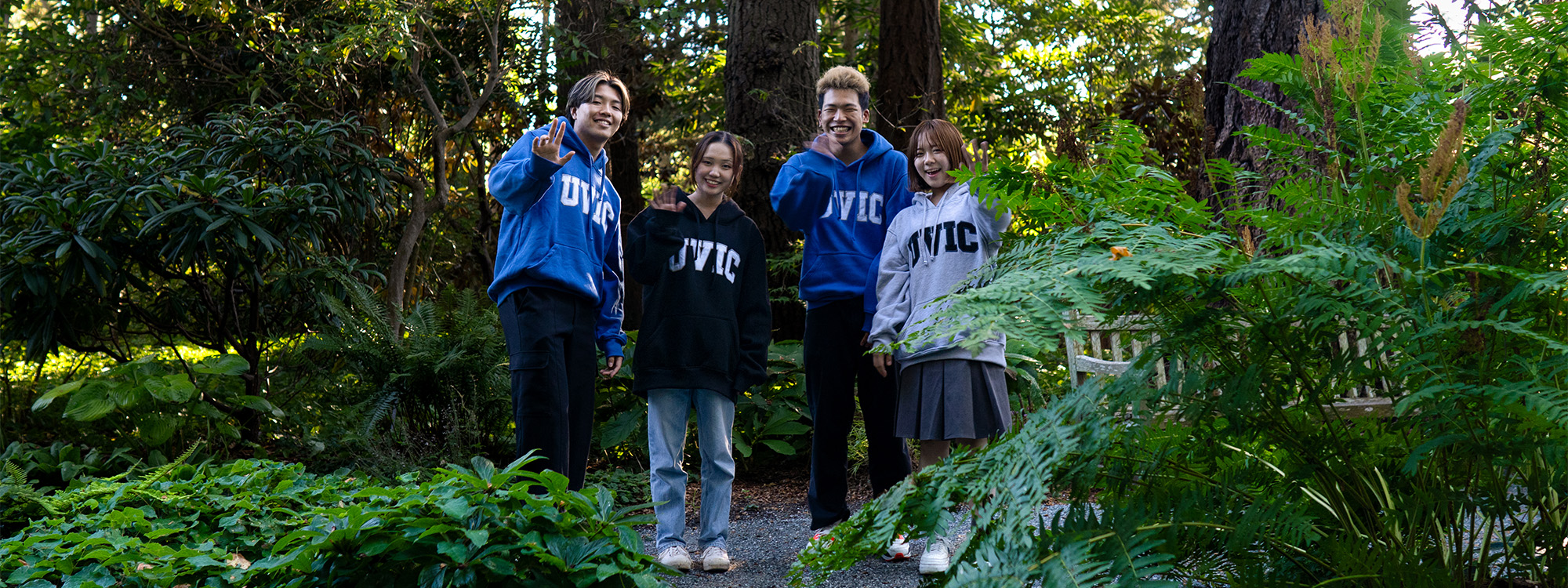Four UVic English Language Centre students walking together through a green forested area on campus, wearing UVic hoodies and smiling.
