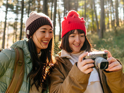 Two women in winter hats enjoying outdoor photography together in a sunlit forest, reviewing photos on a camera—capturing the joy of learning photography through UVic Continuing Studies courses.