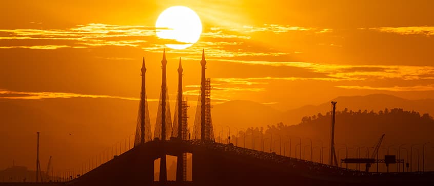 View of the Penang bridge in the background with the sun setting behind it. Golden evening light surrounds the image