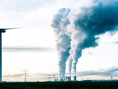 Photo of a green energy windmill juxtaposed with industry smoke towers. 