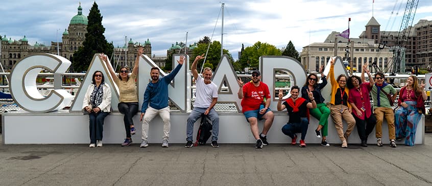 A group of international teachers on a tour of Victoria sitting in front of a sculpture spelling out the word Canada