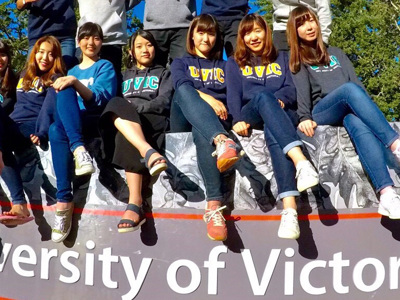 International students sitting on the UVic sign at the entrance to the campus on a sunny day.