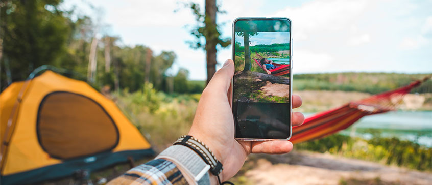 A person holds a smartphone, capturing a camping scene with a hammock and yellow tent near a lake. 