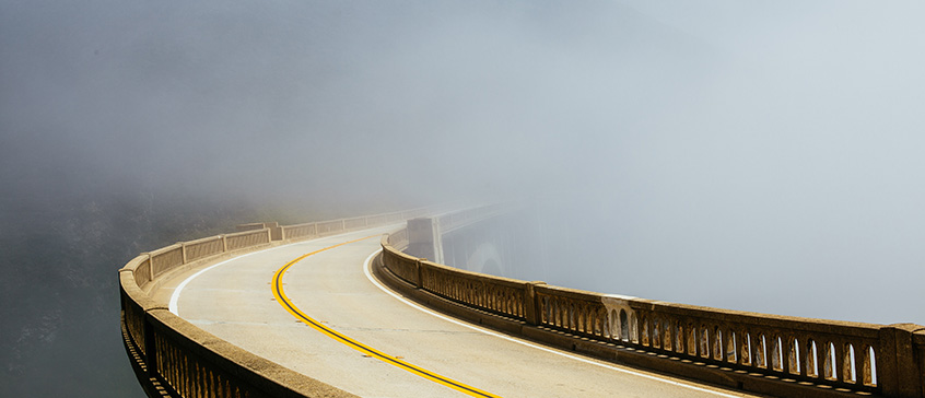 A curved concrete bridge or walkway with yellow center lines winds through dense fog, with safety railings on both sides disappearing into the misty distance, symbolizing navigation through uncertain and challenging circumstances.