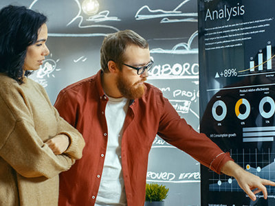 Two individuals reviewing a large digital display with charts, graphs, and analysis metrics in a modern office setting, symbolizing data analytics and business intelligence.