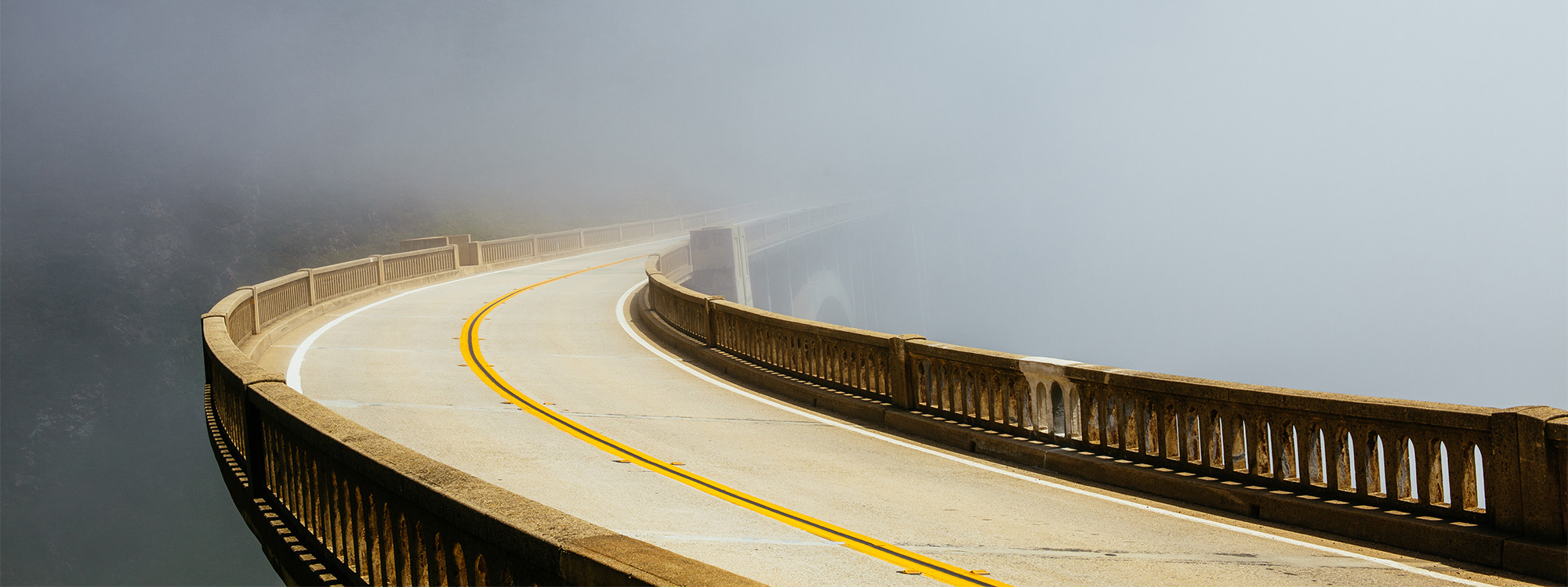 Curving concrete bridge with yellow centre line fading into dense fog.